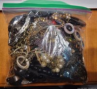 Clear plastic bag filled with assorted costume jewelry items, visible are chain necklaces, clip-on earrings with circular detail, faux pearl pieces, and a wristwatch case without strap.