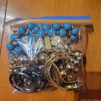 Clear plastic bag filled with assorted costume jewelry including blue bead necklace, white beads, metallic bangles, and other assorted pieces