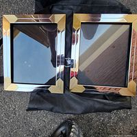 Two rectangular photo frames with dark brown borders and shiny gold ribbed corner accents. Frames have glass fronts and are placed on dark cloth on a paved surface.