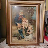 Framed color print showing an elderly man with two children helping to remove his boots, placed against a wall with books visible.