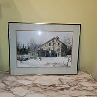 Framed signed print of a winter barn scene on a marble surface.