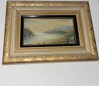 Front view of small framed landscape print with gold ornate frame showing water, trees, and hills.