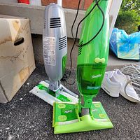 Green Swiffer WetJet floor cleaner next to gray handheld dry vacuum on ground with a cardboard box and shoes nearby.