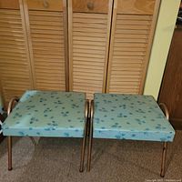 Two matching square turquoise stools, each with brass legs and handles, placed side by side in front of wooden closet doors and carpeted floor.