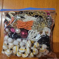 Clear resealable plastic bag filled with assorted bead necklaces in white-yellow striped, orange, black, maroon colors and gold/silver chains.