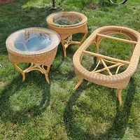 Front view of three light-colored wicker tables on grass: two round with glass tops and one larger oval without glass.