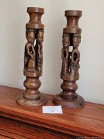 Pair of hand carved antique African wooden lamp bases shown side by side from front angle on wooden surface. Detailed human figures carved on the main shaft.