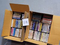 Two open cardboard boxes filled with audio cassette tapes, showing a variety of artists and music compilations from different genres and eras.