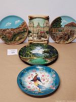 Five items displayed: two round 3D village scene plates, one 3D historic relief plaque, one oval tropical jungle painting plate, one turquoise Asian porcelain bird plate.