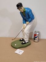 Full front view of the golfer statue showing the putting stance with golf club and ball, positioned next to a Pepsi can for scale.