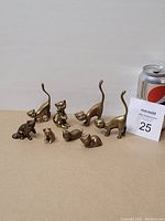 Nine miniature brass cat figurines displayed against a plain background next to a soda can for scale.