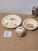 Three Royal Doulton Bunnykins porcelain pieces: divided plate, small bowl, and piggy bank, arranged with a Pepsi can for scale