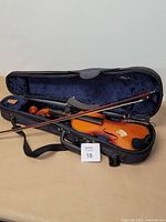Vintage violin inside open black hard case lined with blue velvet; bow lies across the violin and case; violin lacks strings; chinrest attached.