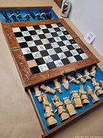 Full view of vintage wooden chess set with open folding carved wooden board showing tile inlay, some tiles have scenic prints, missing one tile, velvet lined drawers with carved chess pieces in light and dark wood.