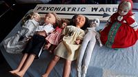 Six vintage dolls of varying materials and sizes arranged horizontally beside a wooden sign with bold black text "PSYCHIATRIC WARD."