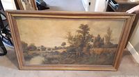 Full view of the large framed landscape painting showing the river, trees and grass with the wooden frame.