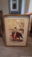 Full view of the framed vintage painting showing the matador and the bull with some visible wear on the wooden frame.