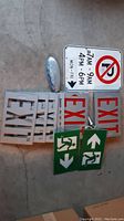 Photo showing multiple Exit signs stacked, No Parking sign and two green emergency exit signs laid on the ground.