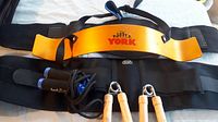 Photo showing 1 vintage yellow/orange York curl support, 2 black back braces, 2 wooden handled hand squeezers, and 1 black and blue skipping rope laid out on a white background