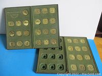 Two green padded presentation cases containing multiple square gold-tone medallions arranged in rows, one case partially empty revealing slots for medallions.