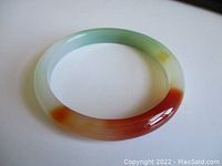 Pale green jade bangle with darker green and red-orange colors lying flat on white surface, showing glossy finish and smooth rounded edges.
