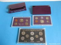 Overview of two USA 1984 proof coin sets in maroon inserts and hard plastic cases, and one Bank of Greece coin set in a black background hard plastic case.
