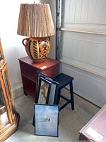 Photo of the corner setup showing the lamp on a small wooden cart, two framed artworks leaning against the cart, and a black saddleback stool next to the cart.