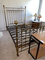 Full view of the metal brass bed frame showing headboard, footboard, side rails, and casters on an indoor carpeted floor.