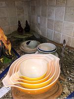 Photo showing four nested yellow Pyrex mixing bowls, ceramic bowls and plates stacked behind, green glass cake stand, red glass cruets, paper towel holder and wooden apple bowl on granite countertop.
