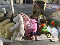 Photo showing assortment of plush toys including white bunny in pink outfit, teddy bear, dolls in box, and other small toys and a children's book.