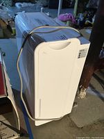 White Home brand dehumidifier standing on basement floor with power cord wrapped on top.