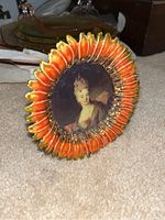 Decorative orange and gold frame with a portrait of a woman, likely from or inspired by a historic painting, resting on carpet.