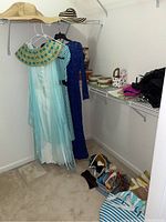 Wide shot showing clothing racks with three dresses, including blue and gold costume dress and blue sequined dress, hats on top shelf, decorative boxes, bags, scarves, and gloves on shelves and floor.