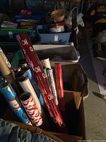Multiple rolls of Christmas wrapping paper stored upright inside a cardboard box, in front of plastic bins containing other holiday decor items.