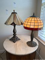 Two metal base table lamps with Tiffany style stained glass shades on a round marble top table near a window.