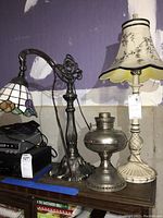 Overall shot showing two electric lamps and one metal oil lamp base on a wooden surface against a purple wall.
