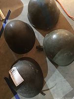 Four military helmets arranged on the floor, showing their exterior front and side views, worn green to dark color finish with one helmet featuring a blue stripe.