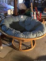 Round rattan papasan chair base with attached large tufted cushion in blue-gray fabric lying on basement floor.