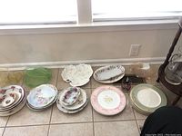 Various ceramic plates and bowls with floral patterns, pastel colors, and some with gold trim, arranged on the floor in front of a window.