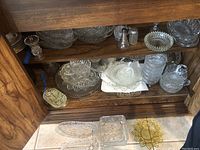 Wooden cabinet shelf with multiple clear cut glass serving bowls, footed dishes, a bud vase, a creamer jug, and round coasters displayed.