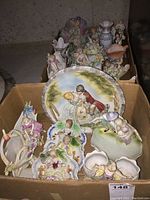 A box containing multiple antique porcelain figurines and wall plaques showing couples, women, and decorative scenes in pastel colors.