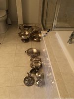 Photo showing the full collection of silver plated serving pieces arranged on the floor near a bathtub and toilet. Items include two chafing dishes, cream sugar set, gravy boat, butter dish, platter, bread bowl, and divided tray.