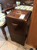 Wooden rectangular waste box showing front and side with wooden handle, surrounded by furniture with patterned cushions.