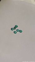 Six small, round cut green emerald stones laid out on neutral background, showing overall size and color.