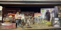 Wide view of shelf with kitchenware assortment, including Pyrex percolator, metal flour sifter, skewers, wine glasses, and boxed items.