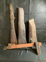 Three leather rifle sling sleeve cases laid out vertically on black background along with the leather belt with attached holster.