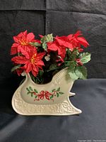 Side view of cream-colored Lenox porcelain holiday sleigh decorated with holly and ribbon motif, filled with red faux poinsettia flowers and green leaves.
