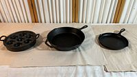 Three cast iron cookware items displayed on a beige cloth: a round muffin pan on the left, a large skillet in the center, and a smaller skillet on the right.