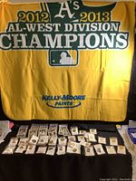 Full view of yellow and green fleece blanket with text for 2012-2013 AL-West Division Champions Oakland Athletics, displayed behind an assortment of individually bagged and carded commemorative pins.
