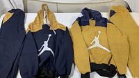 Front view showing two hooded sweatshirts side by side with basketball player logos visible and matching jogger pants in navy and mustard colors.